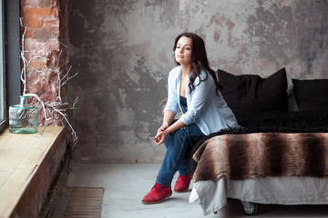 Sensual woman with dark hair in blue shirt and jeans sitting on a bed at home. Loft style interior