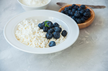 Organic Farming Cottage cheese in a plate sour cream and blueberries on a white stone background. Copy space.