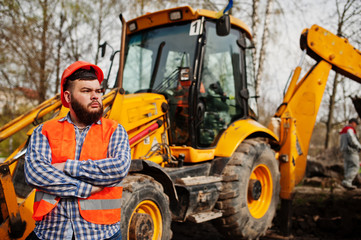 Brutal beard worker man suit construction worker in safety orange helmet against traktor.