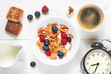 Healthy breakfast on white table. Top view of bowl of cornflakes with berries and nut, black coffee, milk, cinnamon cookie and alarm clock. 