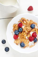 Vertical top view of bowl of cornflakes with raspberry, blueberry, walnut and fresh milk on white wooden table. 