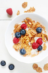 Vertical top view of bowl of cornflakes with milk, raspberry, blueberry and walnut on white wooden table. 
