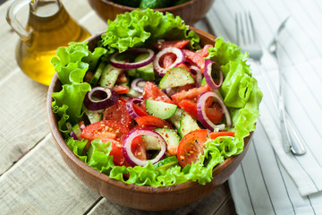 Rustic salad of fresh tomatoes, cucumbers, red onions and lettuce, dressed with olive oil and ground pepper in a wooden bowl. Top view