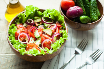 Rustic salad of fresh tomatoes, cucumbers, red onions and lettuce, dressed with olive oil and ground pepper in a wooden bowl. Top view