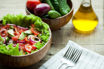 Rustic salad of fresh tomatoes, cucumbers, red onions and lettuce, dressed with olive oil and ground pepper in a wooden bowl. Top view