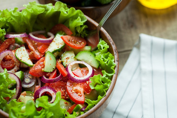 Rustic salad of fresh tomatoes, cucumbers, red onions and lettuce, dressed with olive oil and ground pepper in a wooden bowl. Top view