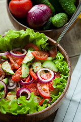 Rustic salad of fresh tomatoes, cucumbers, red onions and lettuce, dressed with olive oil and ground pepper in a wooden bowl. Top view