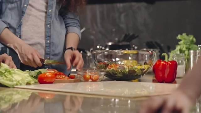 Tracking shot of man and woman chopping salad and tomatoes for dinner on kitchen counter; man drinking red wine