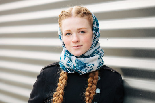 Young beautiful fashionable redhead woman with braids hairdo in blue white headcraft stylish denim black trench jacket posing outdoors against urban style background of metal strips