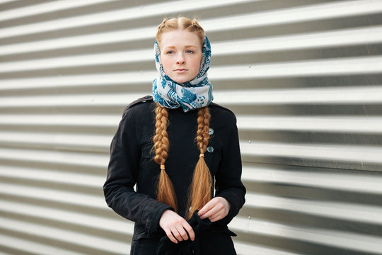 Young beautiful fashionable redhead woman with braids hairdo in blue white headcraft stylish denim black trench jacket posing outdoors against urban style background of metal strips