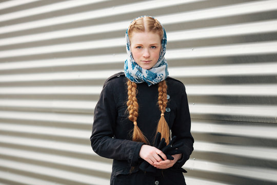 Young beautiful fashionable redhead woman with braids hairdo in blue white headcraft stylish denim black trench jacket posing outdoors against urban style background of metal strips