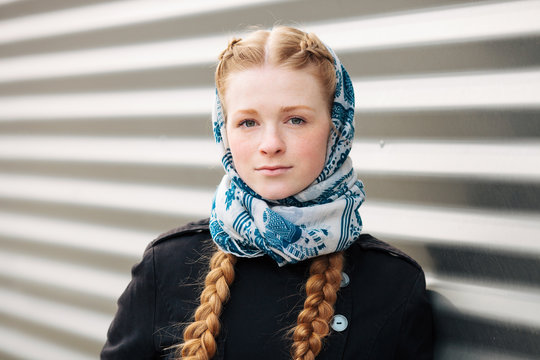 Young beautiful fashionable redhead woman with braids hairdo in blue white headcraft stylish denim black trench jacket posing outdoors against urban style background of metal strips