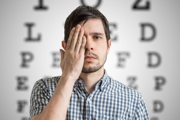 Young man is covering his face with hand and checking his vision. Chart for eye sight testing in background.