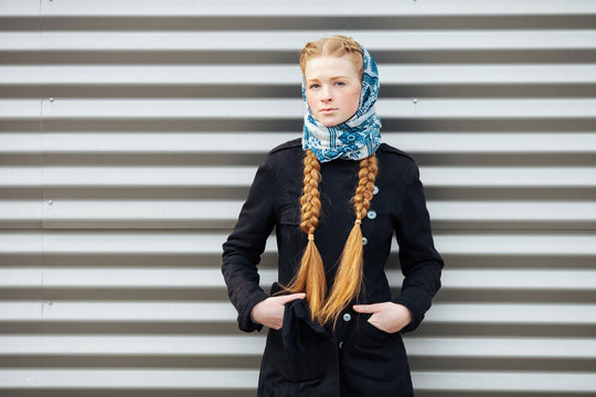 Young beautiful fashionable redhead woman with braids hairdo in blue white headcraft stylish denim black trench jacket posing outdoors against urban style background of metal strips