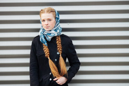 Young beautiful fashionable redhead woman with braids hairdo in blue white headcraft stylish denim black trench jacket posing outdoors against urban style background of metal strips