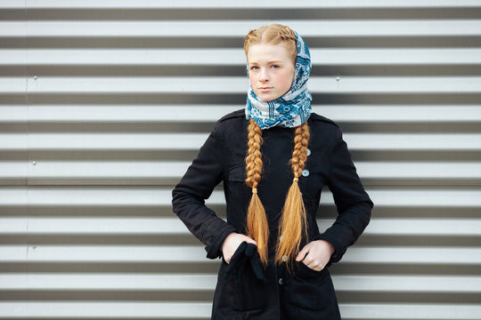 Young beautiful fashionable redhead woman with braids hairdo in blue white headcraft stylish denim black trench jacket posing outdoors against urban style background of metal strips