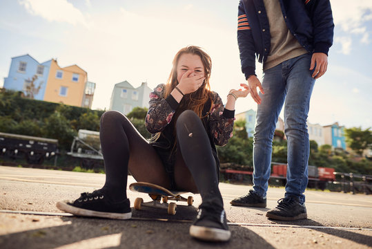 Two friends fooling around outdoors, young woman sitting on skateboard, laughing, low section, Bristol, UK