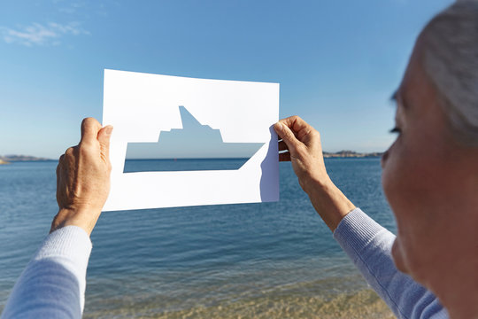 Senior woman standing facing the sea, holding out paper with cut-out of boat