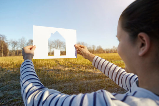 Woman standing in rural setting, holding out paper with cut-out of house