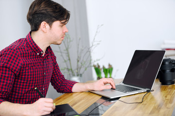 Young cute guy working in an office with a laptop and editing photos using a graphic tablet