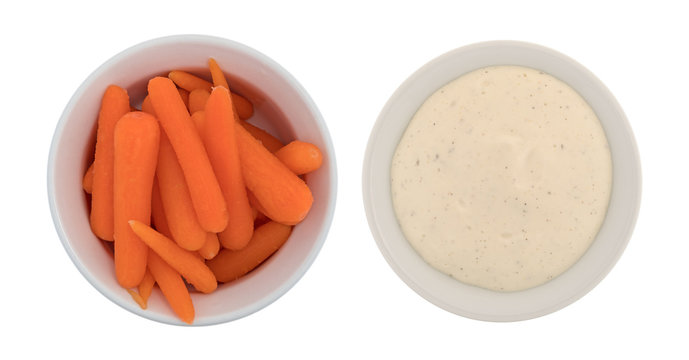 Top View Of Peeled Small Carrots In A Bowl With Ranch Dressing To The Side Isolated On A White Background.