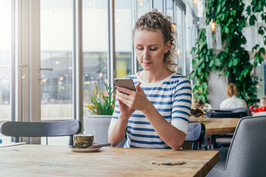 Young business woman in striped T-shirt is sitting at table in cafe near window and is using smartphone.