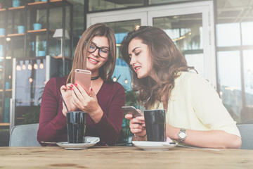 Two young cheerful women sit at table in cafe and use smartphone.