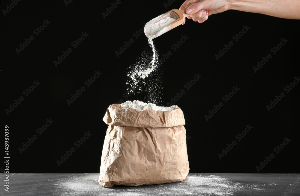 Sticker bag of flour and female hand with scoop on dark background