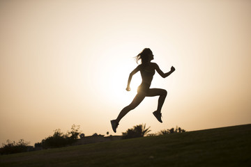 Fototapeta na wymiar Dark silhouette of an athletic woman leaping as she jogs with a sunset in the background 