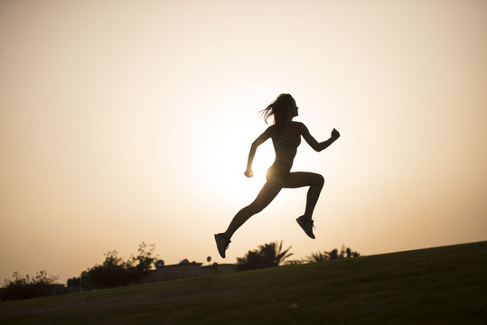 Dark silhouette of an athletic woman leaping as she jogs with a sunset in the background  