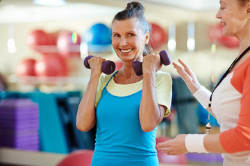 Smiley elderly female looking at camera while instructor helping her to do exercise