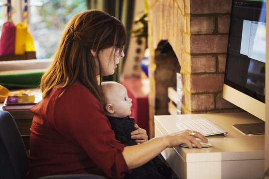 A woman seated with a baby on her lap using her computer, both watching the screen. 