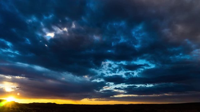 A static sunset timelapse with dramatic clouds against a moody dark blue and orange sky with the sun breaking thorough the clouds just before it sets and a sickle moon peaking thru