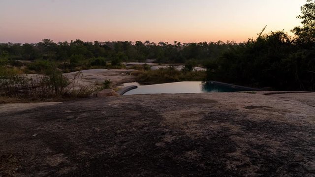 A static timelapse shot from a luxurious African lodge with a private swimming pool at dawn, just before sunrise framed by granite rocks in the foreground