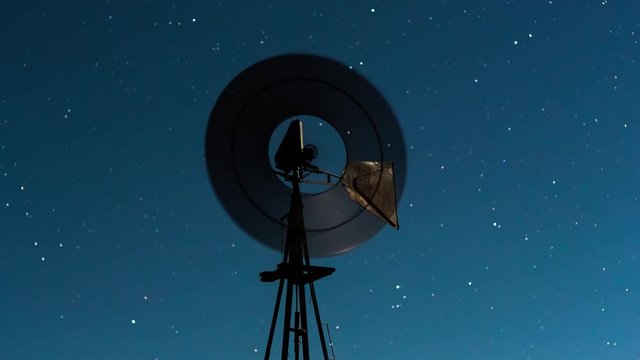 A linear push-in nighttime timelapse of a silhouette windmill blowing in the wind, highlighted by the moon shine on the metal tail, with slow moving stars against a blue moonlight night sky