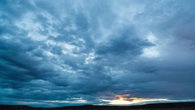 An early morning timelapse before sunrise with dark blue moody clouds rolling through while tilting down to a semi silhouette landscape and an orange glow in the sky
