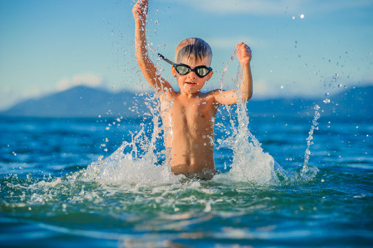 Waist portrait on a tropical beach: handsome boy in goggles jumps up and landed in the water, a lot of splashes fly apart - ready to fly like a bird, having fun