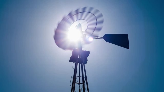 Static daytime timelapse of a silhouette Windmill frantically blowing in the wind against a blue sky and the bright sun