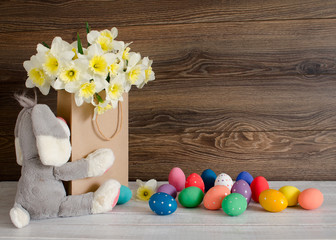Fototapeta na wymiar Colorful Easter eggs with lovely bouquet of narcissus in paper bag and funny bunny on wood background
