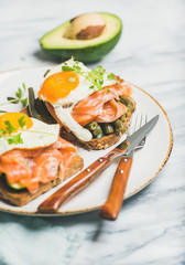 Healthy breakfast sandwiches. Salmon, avocado, fried egg, sauted green beans and fresh sprouts sandwiches in white plate over marble background, selective focus. Clean eating, dieting food concept