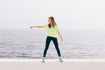 Beautiful slim girl in green sportswear shows the direction of the hand on the background of the sea