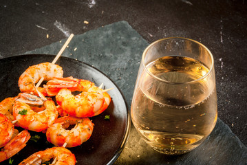 Seafood. Shellfish. Fried grilled shrimp prawns on wooden skewers, with sauce of olive oil and fresh herbs, glass of wine. Snack, appetizer. On black concrete table, copy space