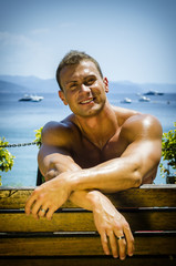 Handsome Muscular Shirtless Hunk Man Outdoor at Seaside Looking at Camera, Sitting on Bench under the Sun