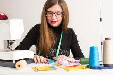 needlework in the workshop of a young woman, a tailor - pretty tailor in glasses at work with pieces of colored cloth on the table with threads, fabrics, needles and sewing machine. Hobby concept.