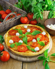 Italian pizza with mozzarella, cheese, and basil leaves on rough rustic rural planks table. Wicker wooden basket with fresh raw organic tomatoes.
