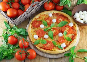 Italian pizza with mozzarella, cheese, and basil leaves on rough rustic rural planks table. Wicker wooden basket with fresh raw organic tomatoes. Horizontal,  top view..