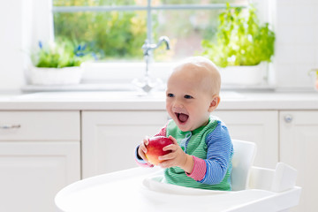 Baby boy eating apple in white kitchen at home