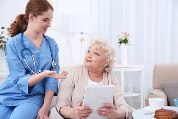 Nurse helping elderly woman to work on tablet pc in light room