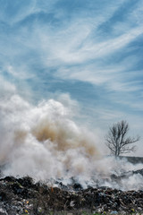 Fire at landfill with white smoke in sunny weather and clouded sky.