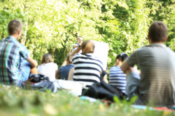 A group of young students in the park. View of a man gesticulating with his hands, standing against a defocused group of people sitting in front of him on the grass.
